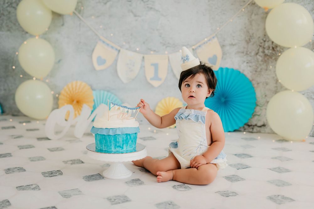 Foto del primer añito de una niña con tarta y peto en tonos amarillos y azules en su sesión en estudio.