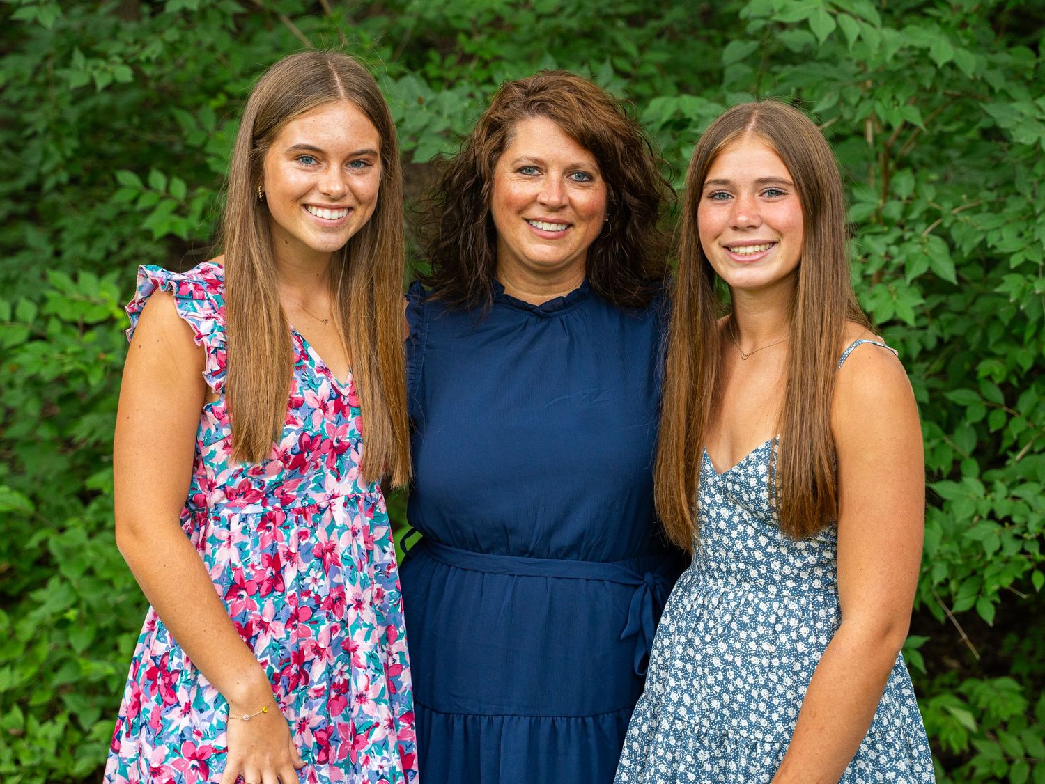 Three people in summer dresses pose together for outdoor family photo with greenery background.