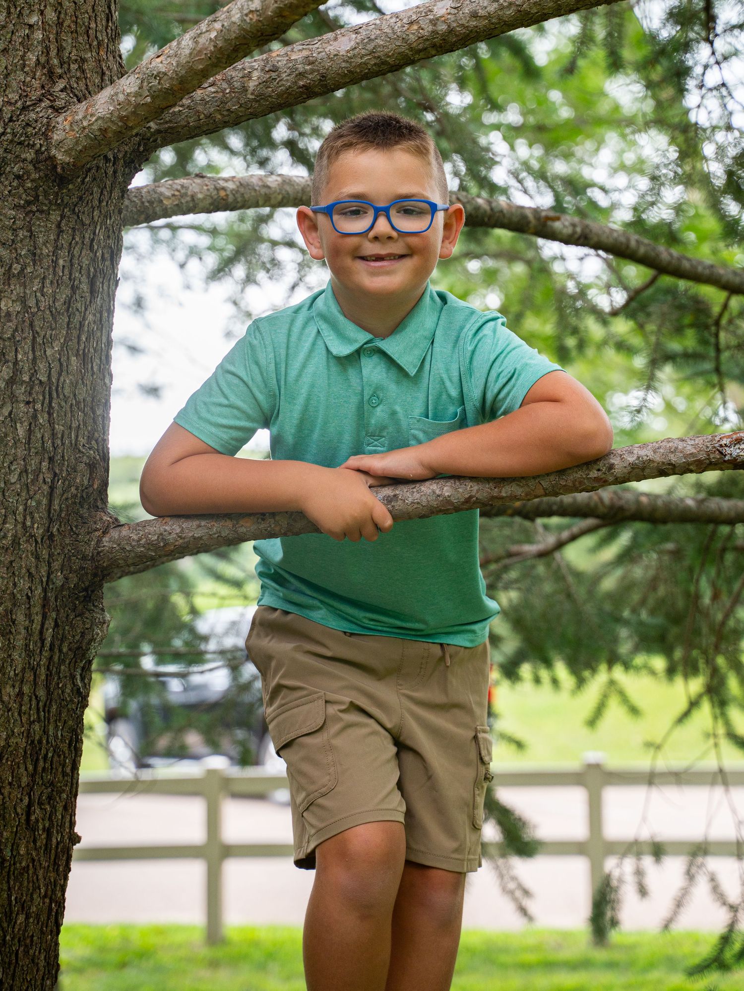 Young person in teal polo shirt and khaki shorts posing playfully in tree.