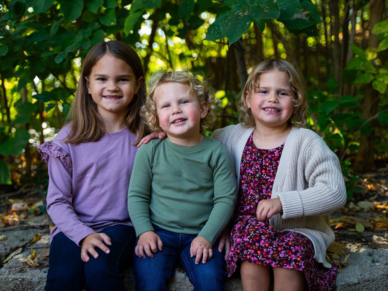 Three young children sitting together outdoors wearing purple, green and floral outfits.