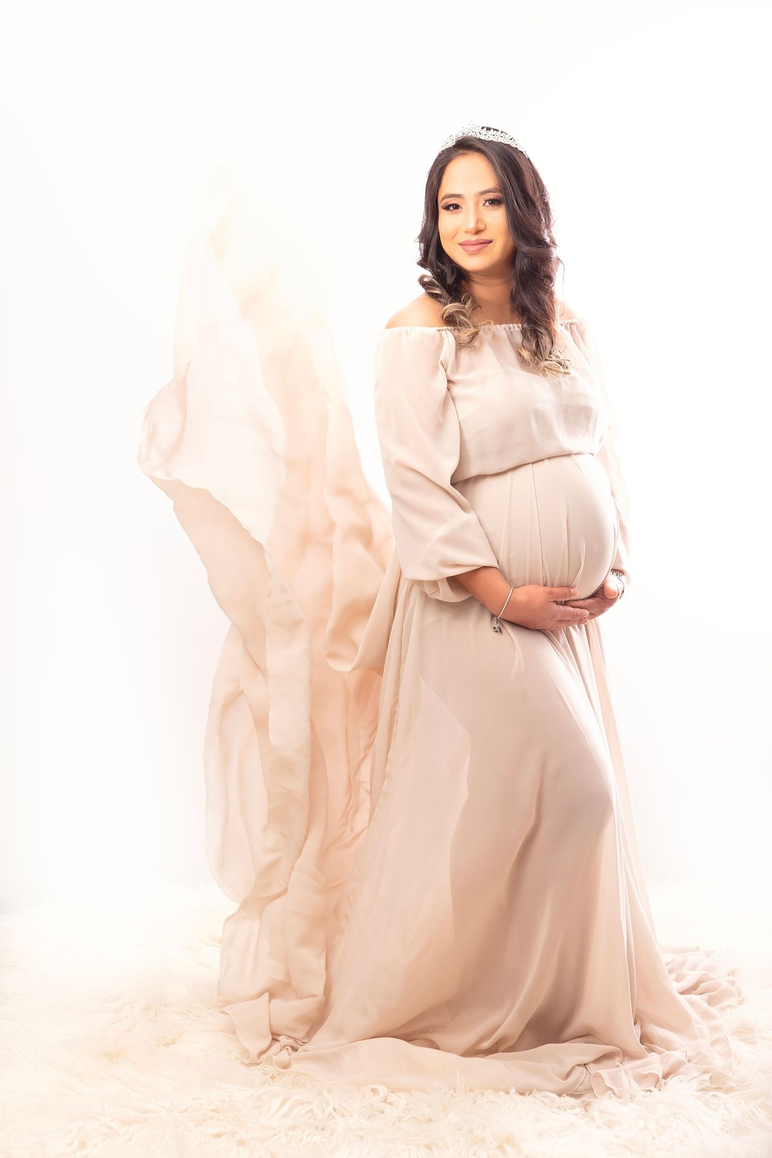 Elegant pregnant woman in flowing beige gown with billowing fabric on white studio background.