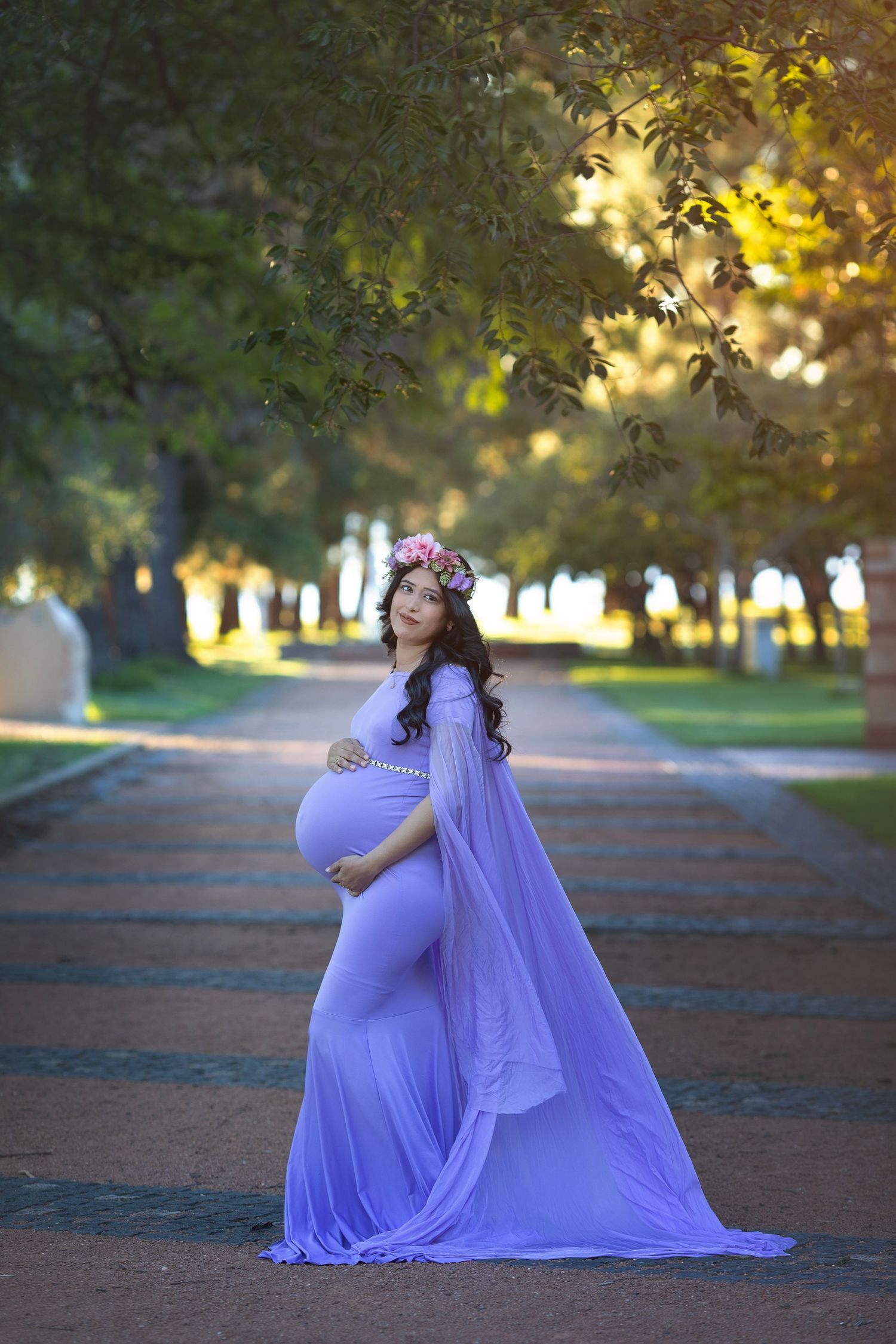 Pregnant woman in lavender gown with floral crown standing on a tree-lined path in a park.