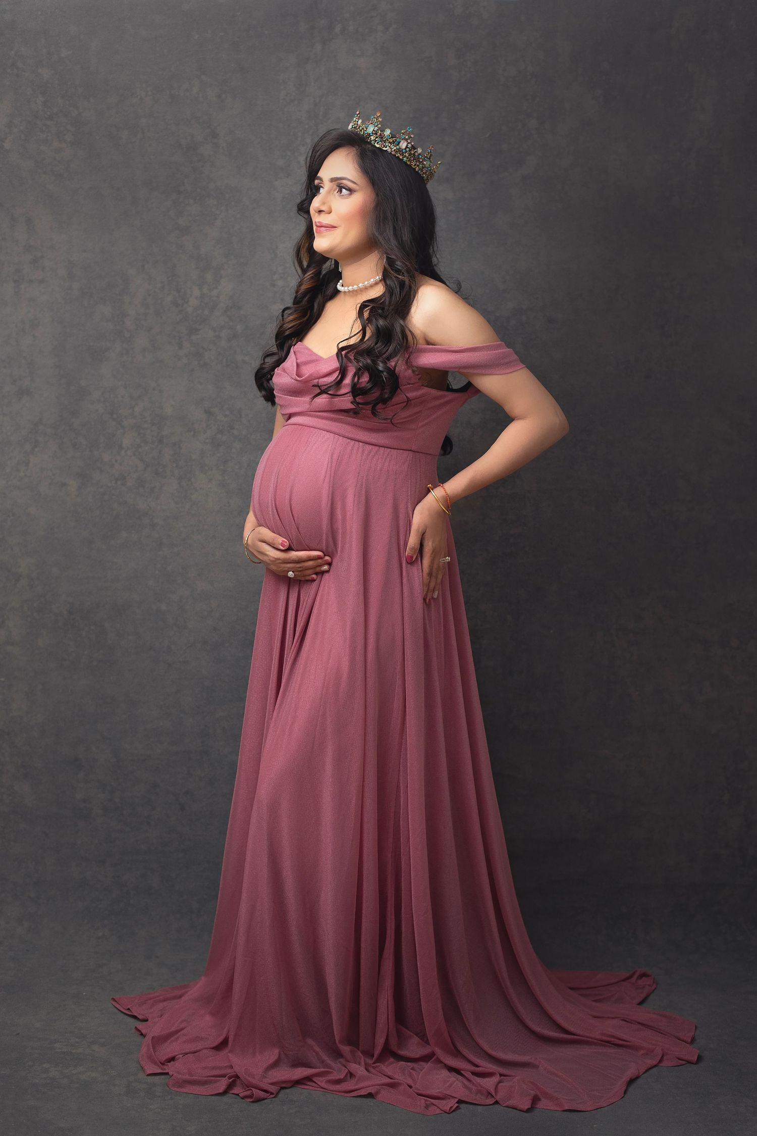 Pregnant woman in dusty rose off-shoulder gown wearing crown posing on gray studio backdrop.
