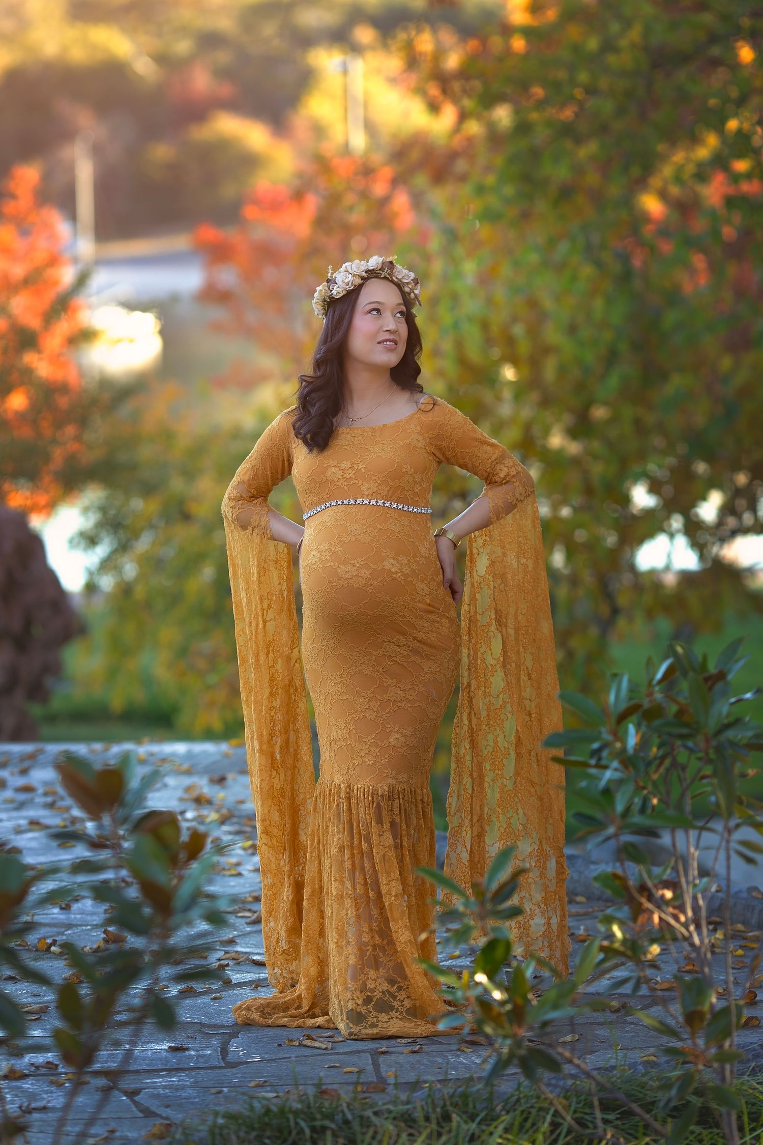 Pregnant woman in golden lace gown with floral crown posing in autumn garden at sunset.