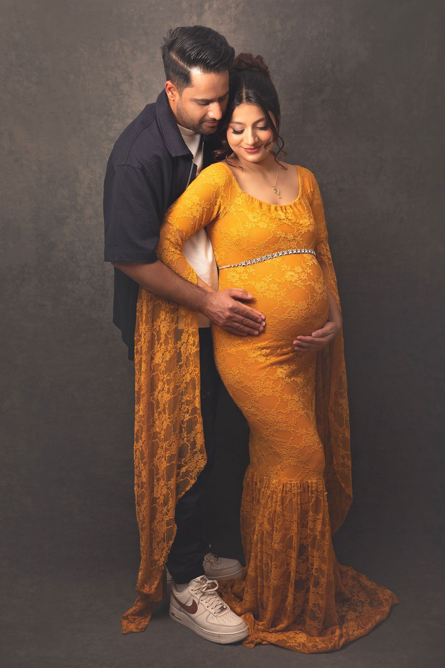 Couple in studio, pregnant woman in gold lace gown with partner embracing her from behind.