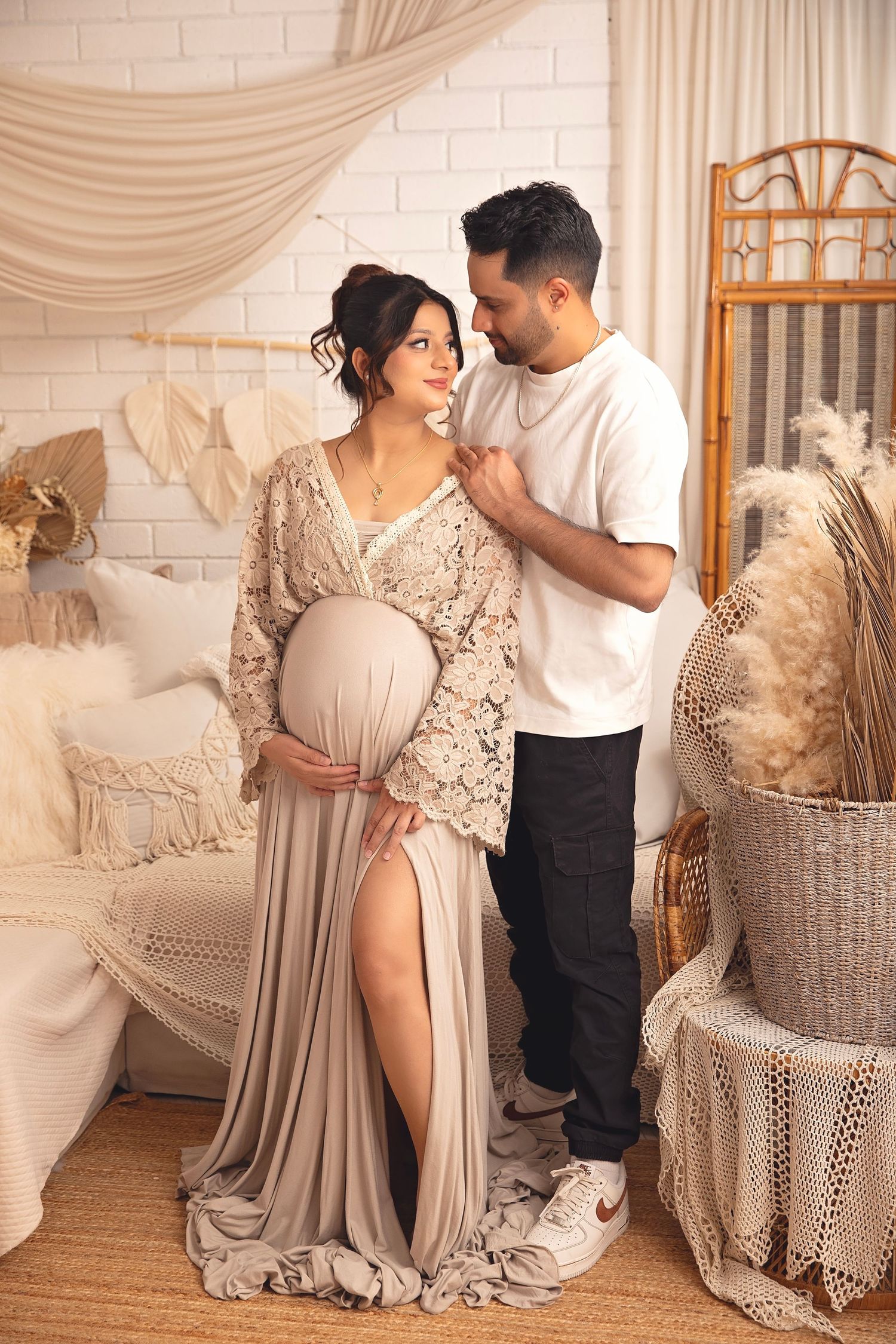Couple in boho studio setting, pregnant woman in lace gown embraced by partner.