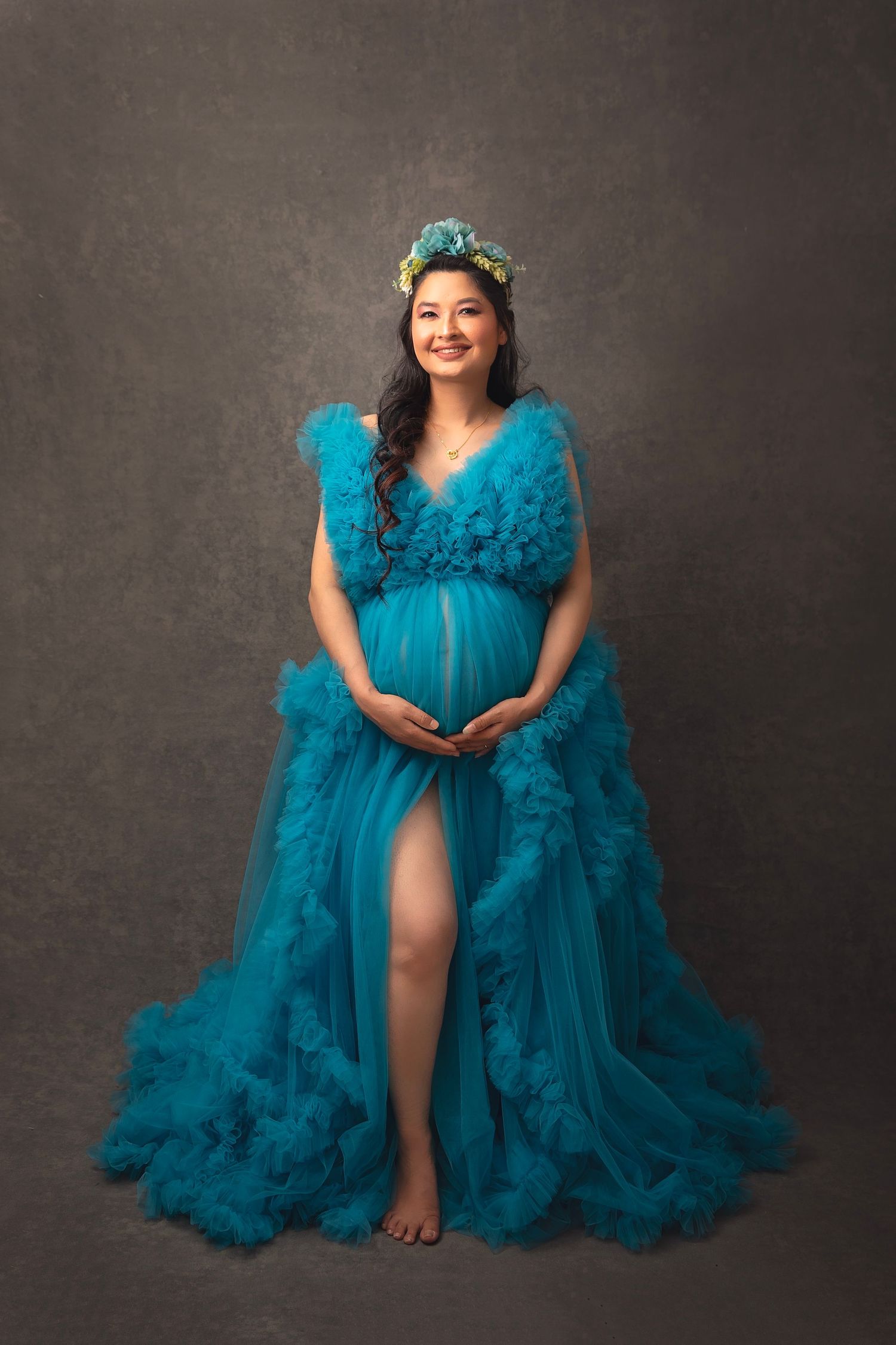 Smiling pregnant woman in teal ruffled tulle gown with floral crown on studio backdrop.