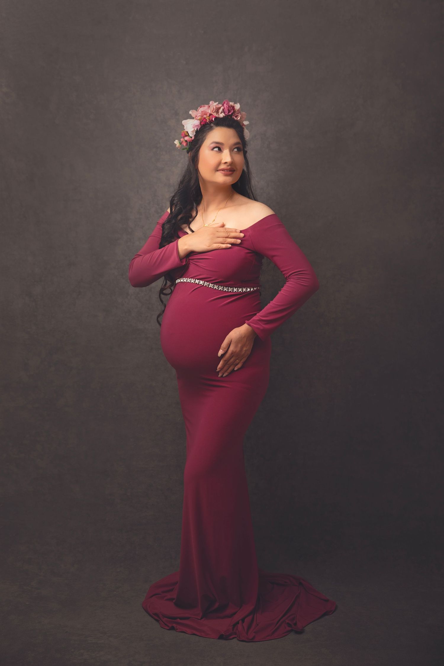 Pregnant woman in burgundy off-shoulder gown with floral crown posing on gray studio backdrop.
