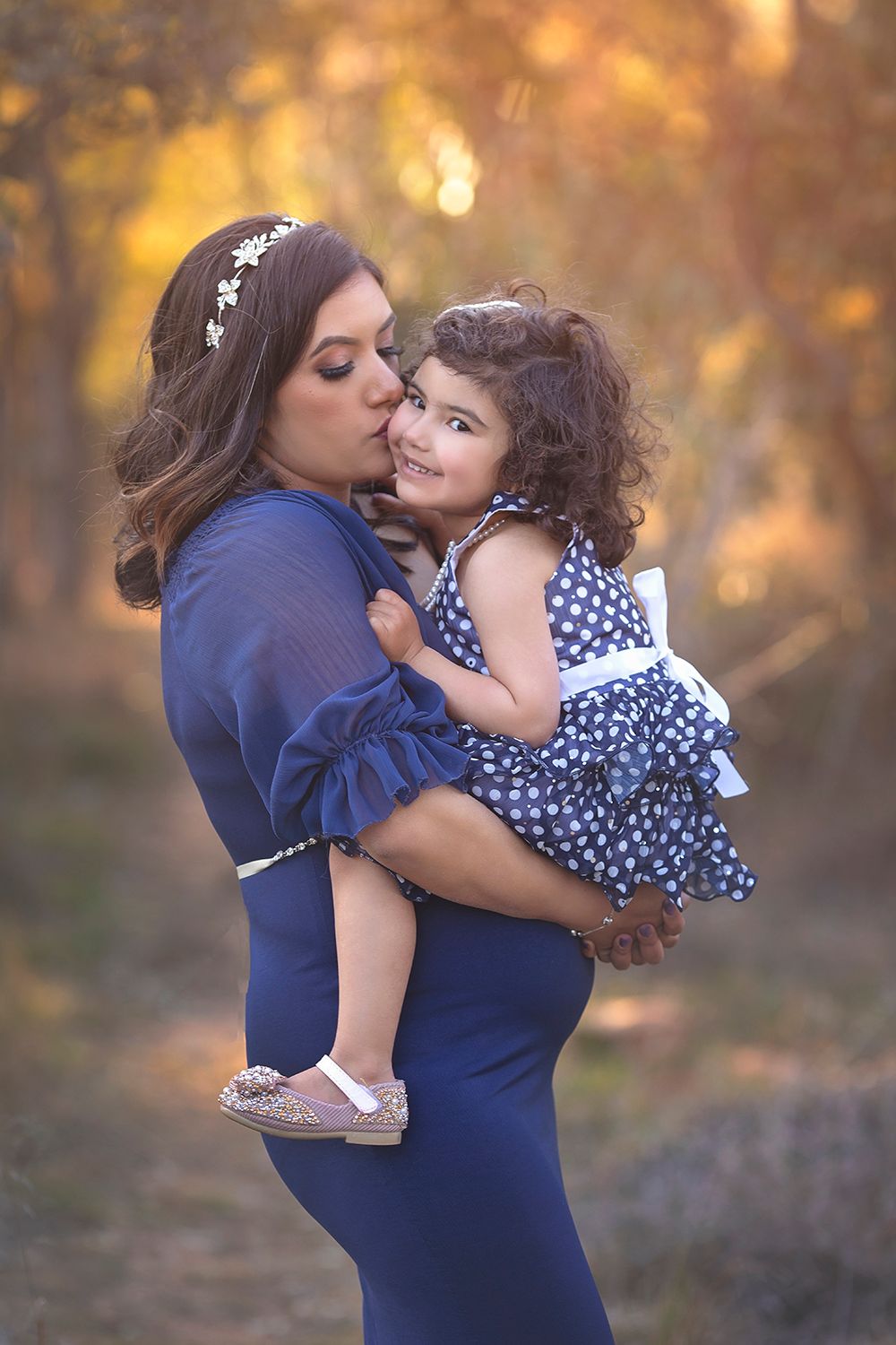 A loving mother in a navy dress kisses her curly-haired toddler daughter in a golden autumn forest.