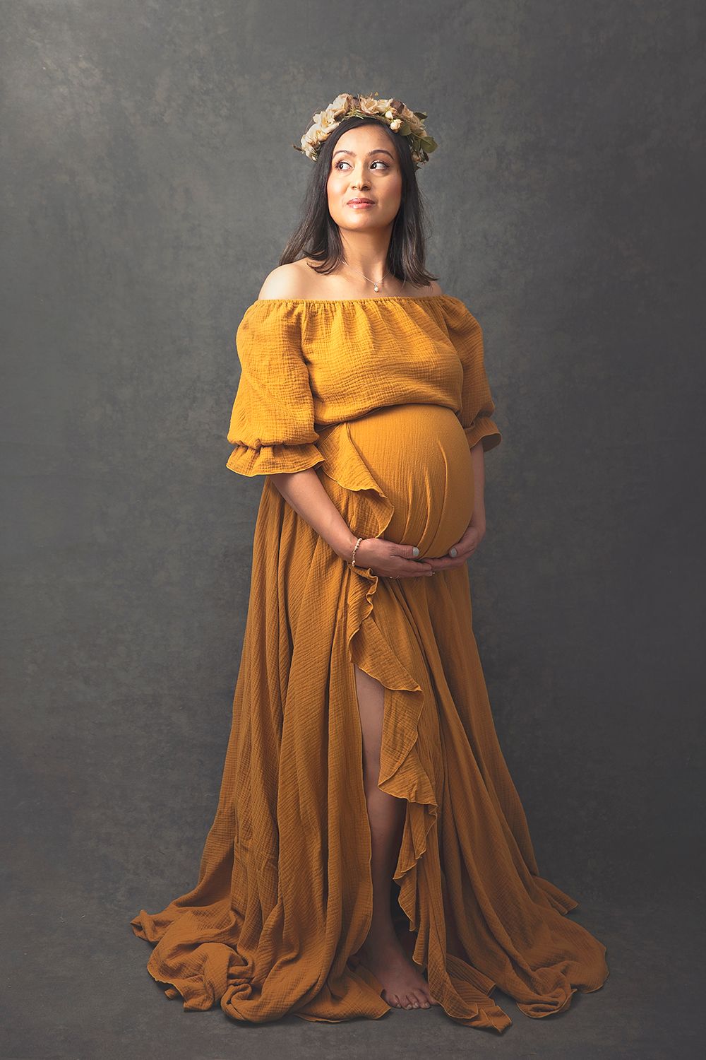 Pregnant woman in a golden off-shoulder maternity gown with a floral crown posing gracefully indoors.