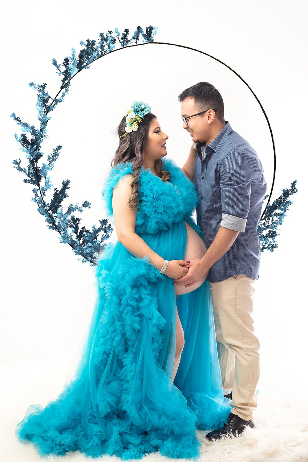 Pregnant woman in teal tulle gown holds hands with partner before floral hoop backdrop.