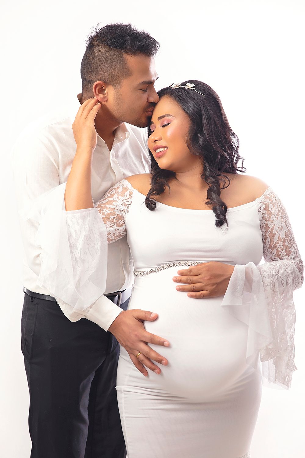 Couple shares tender moment, man kisses pregnant woman in elegant white lace maternity dress.