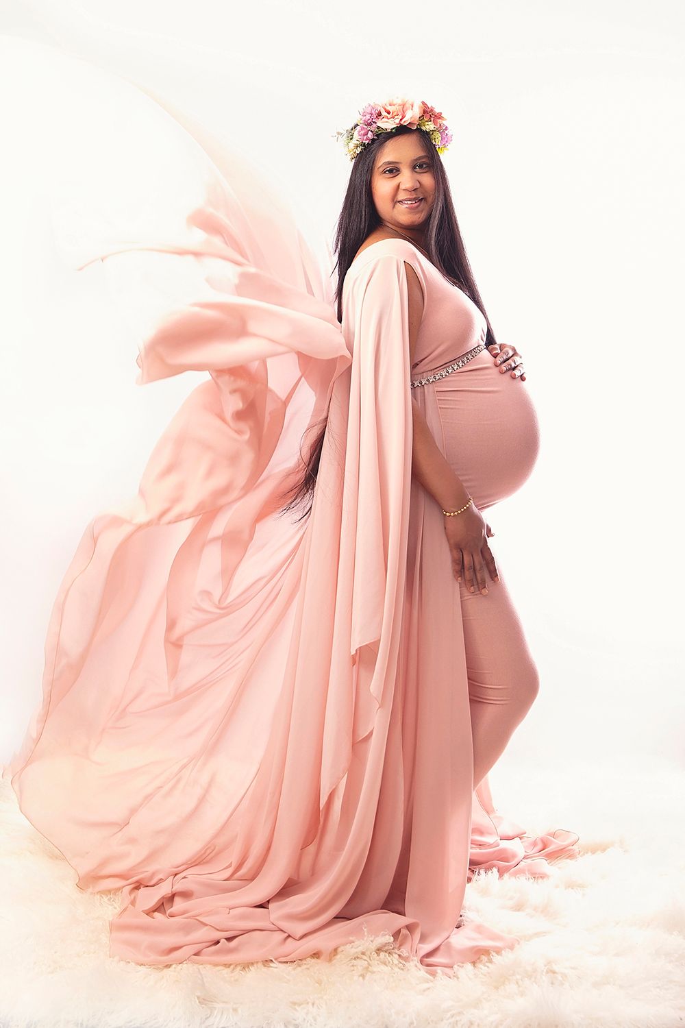 Pregnant woman in flowing pink gown with floral crown poses gracefully in studio.