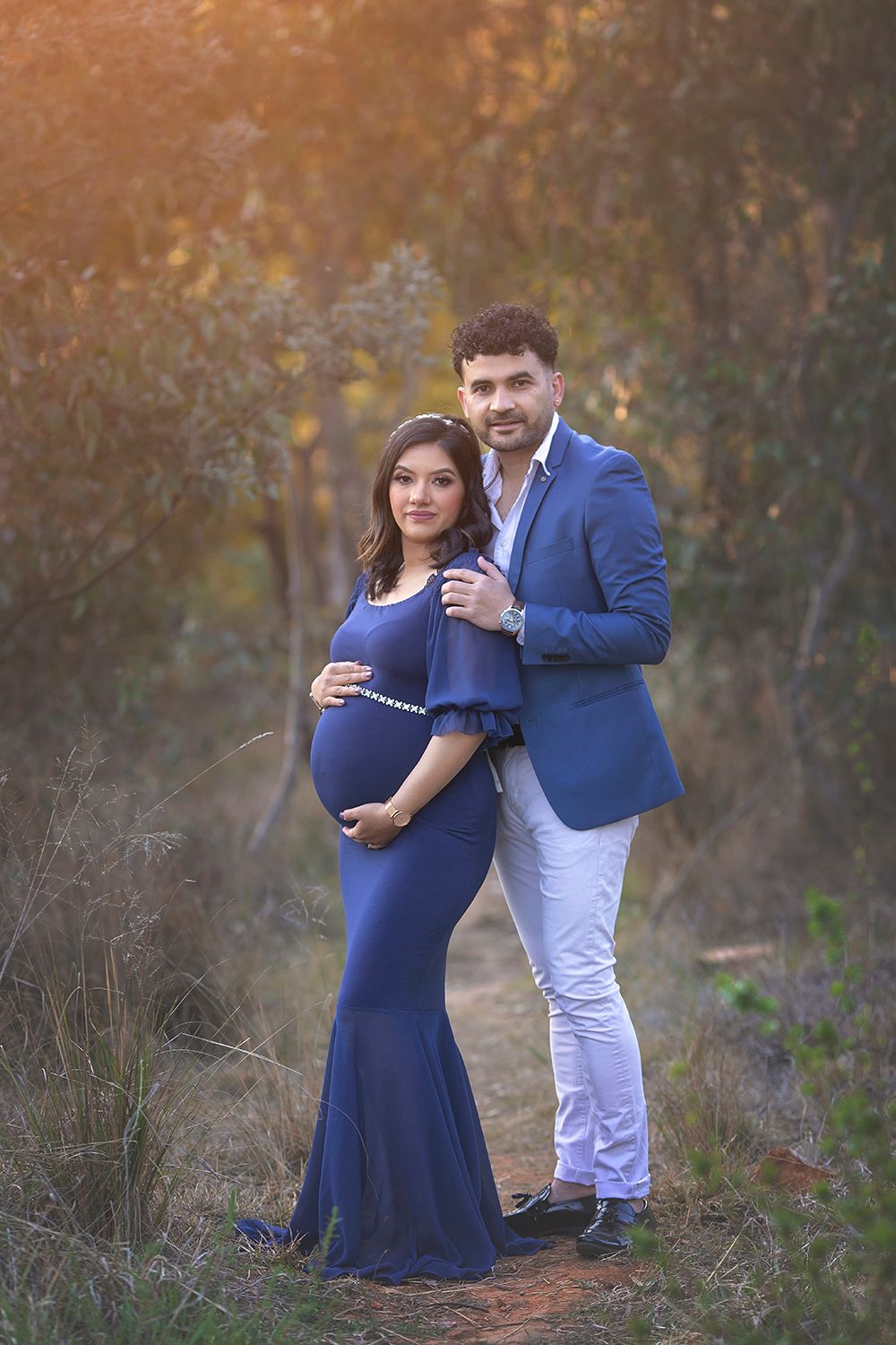 Pregnant couple poses elegantly outdoors at sunset, woman in navy blue maternity gown.