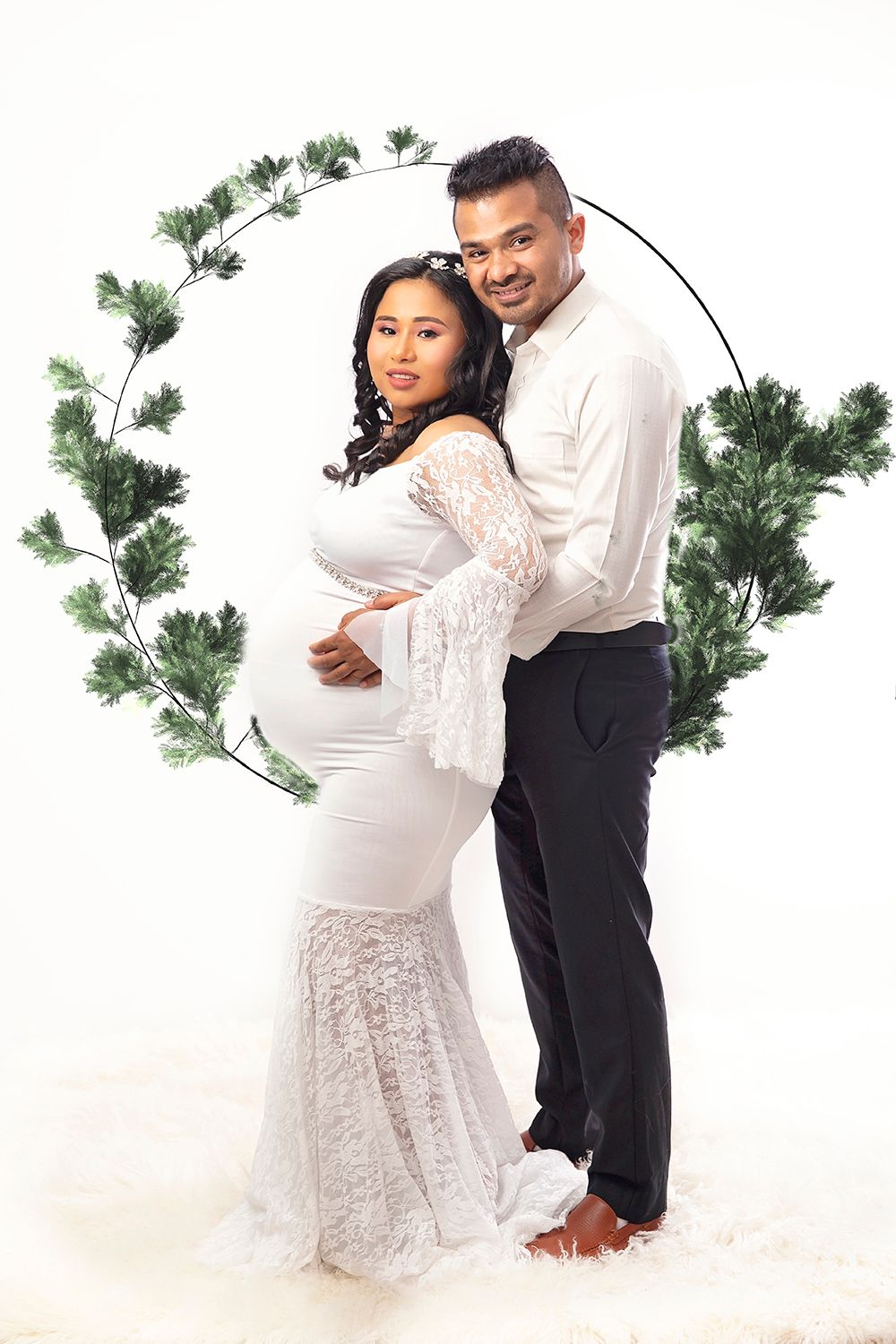 Couple poses before green wreath backdrop, pregnant woman in white lace maternity gown.