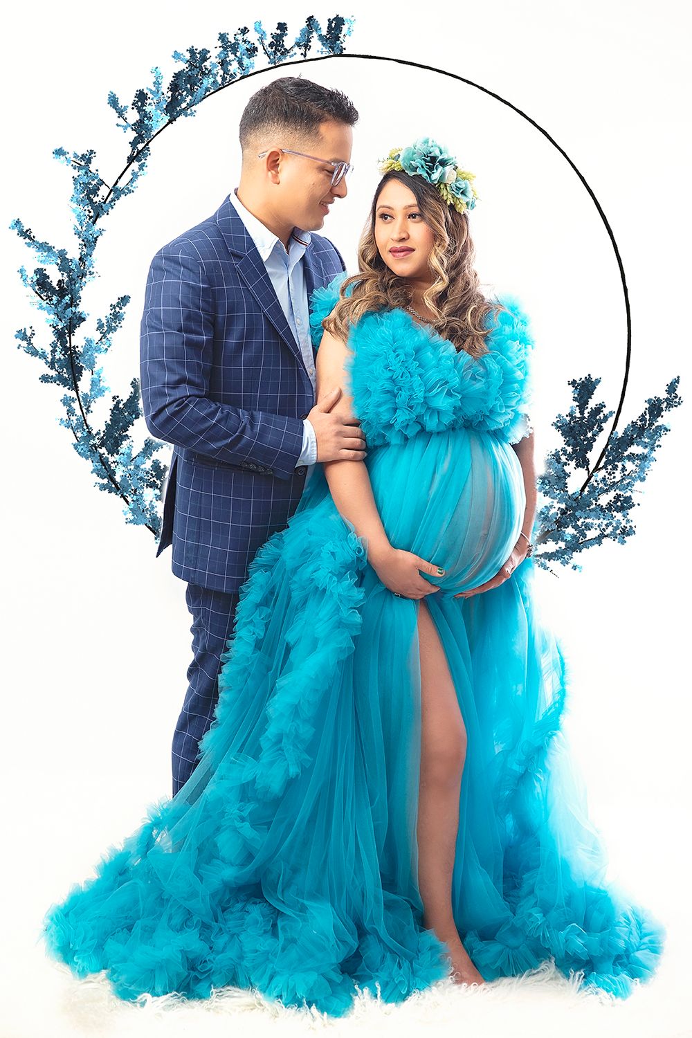 Expecting couple in coordinating blue outfits poses before decorative teal floral hoop backdrop.