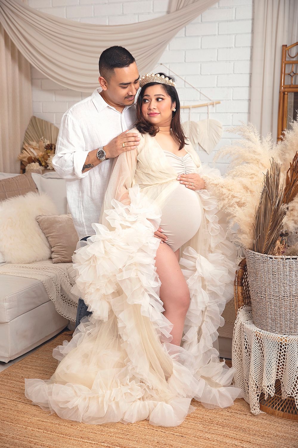 Couple in boho studio setting, pregnant woman in cream ruffled maternity gown with crown.