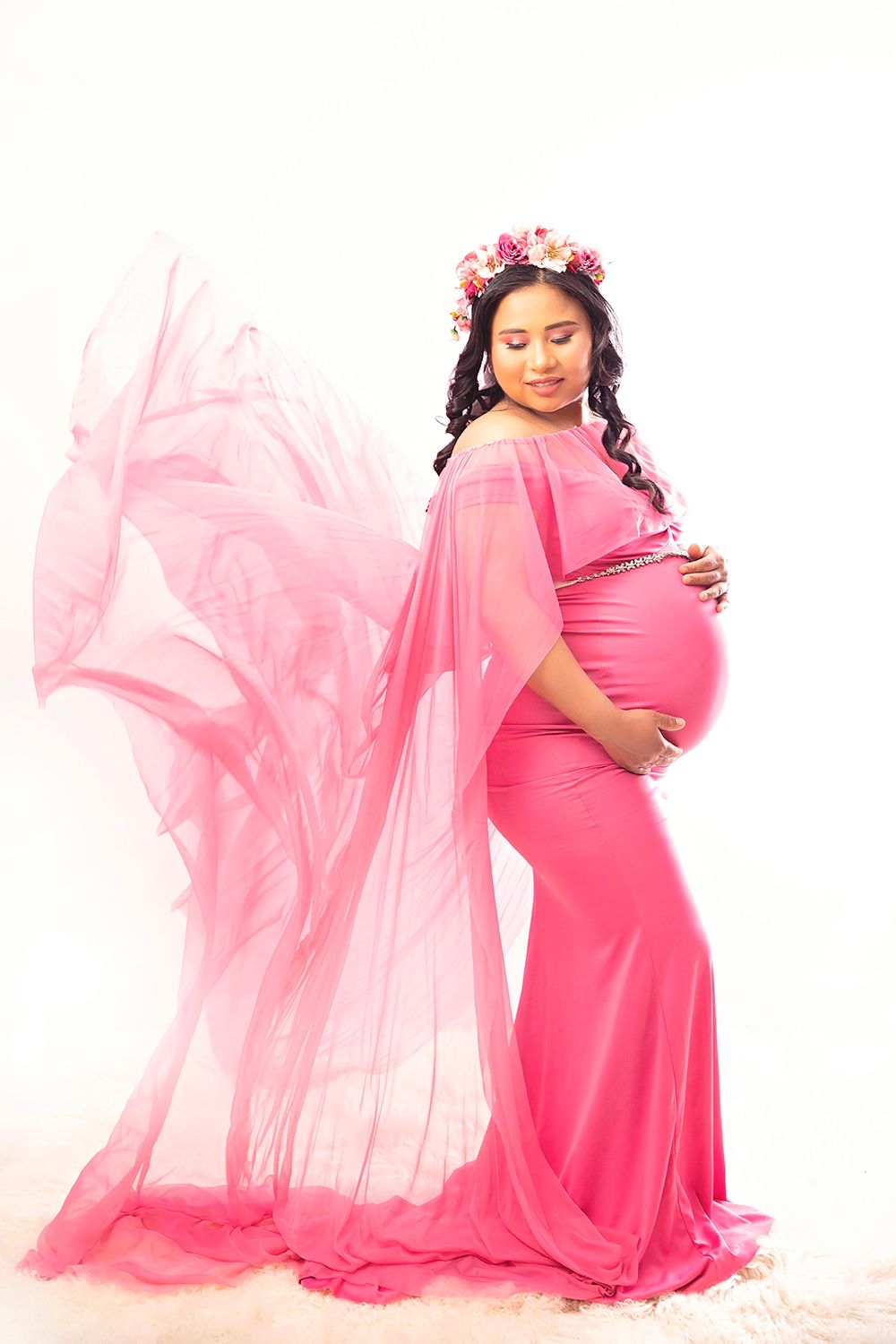 Pregnant woman in vibrant pink gown with floral crown poses gracefully in bright studio.