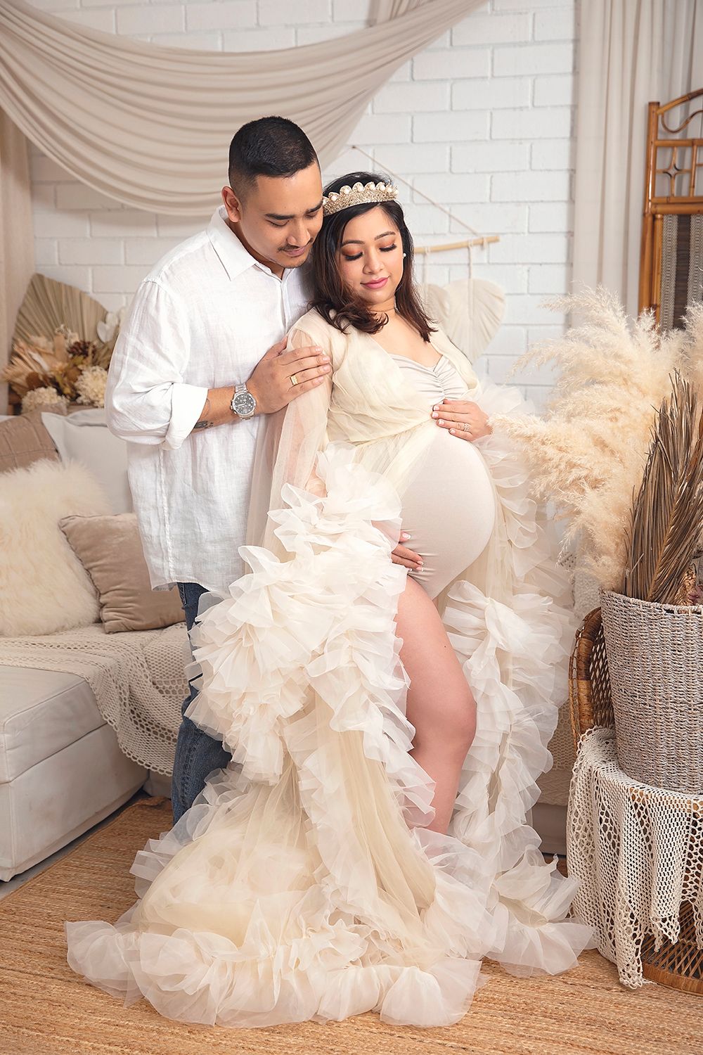 Couple in boho studio, man in white shirt embraces pregnant woman in ruffled cream tulle gown.