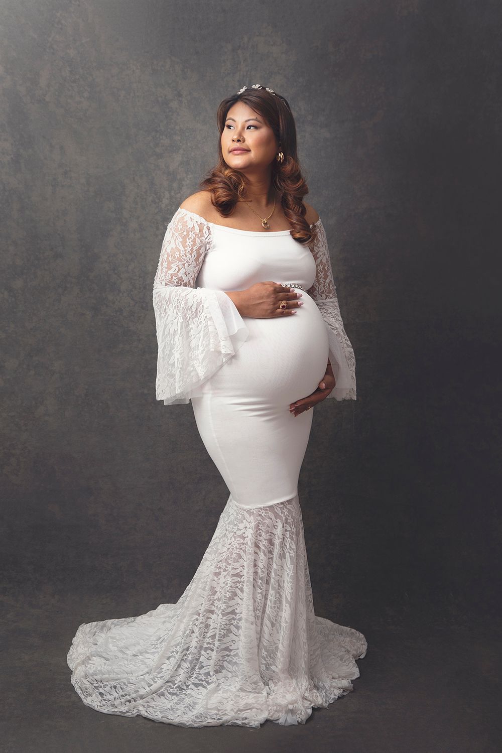 Pregnant woman in elegant white lace off-shoulder mermaid gown poses against gray studio backdrop.