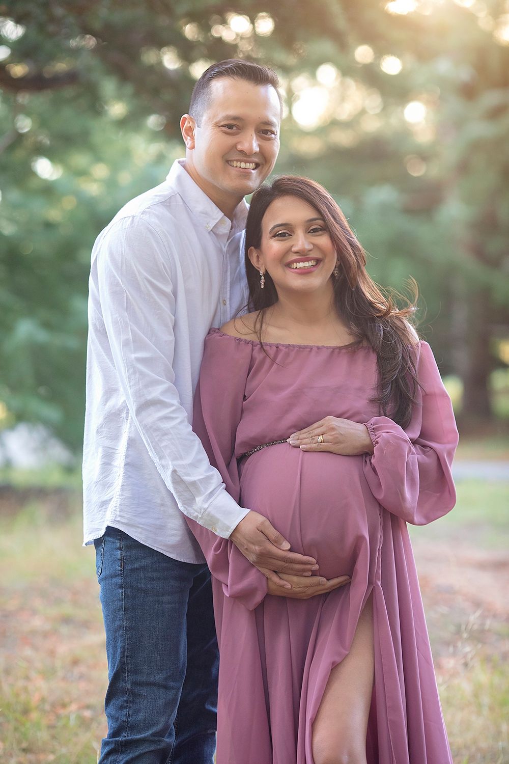 Smiling couple outdoors, pregnant woman in mauve dress held by partner among sunlit trees.