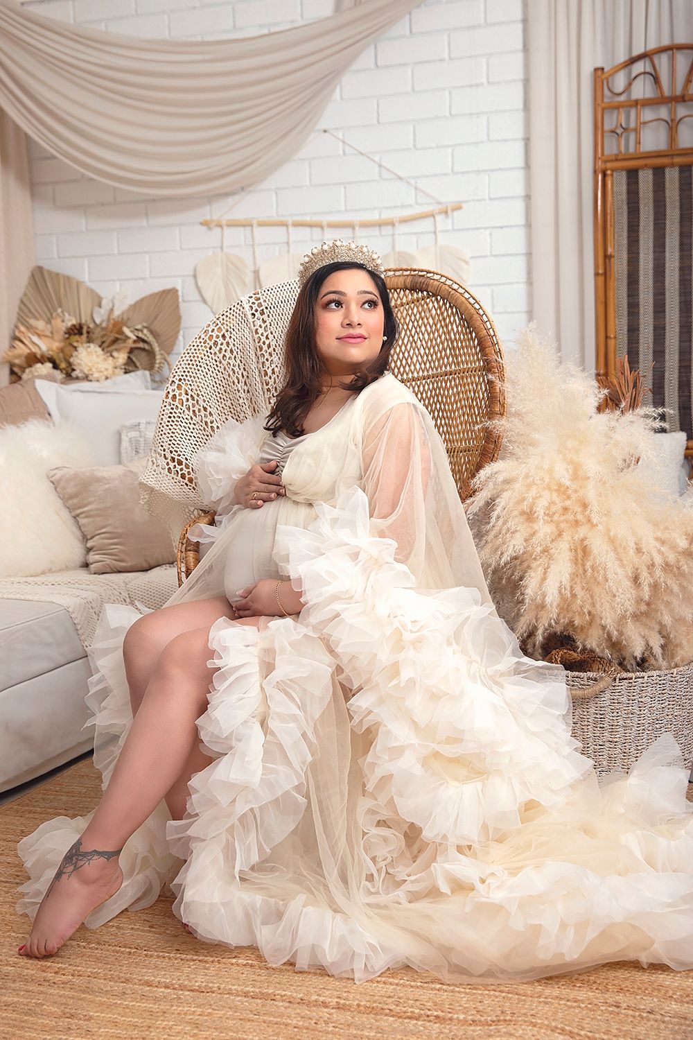Pregnant woman in cream ruffled tulle gown sits in peacock chair in boho-styled studio.