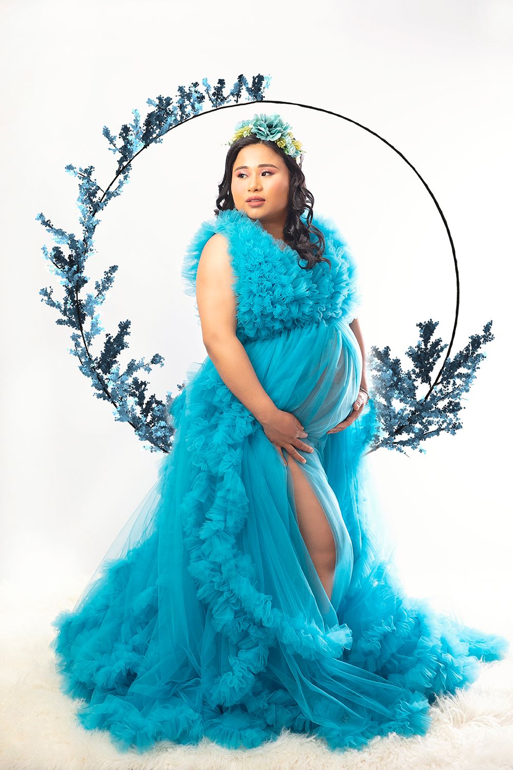 Pregnant woman in turquoise ruffle gown with floral crown poses before circular blue branch backdrop.