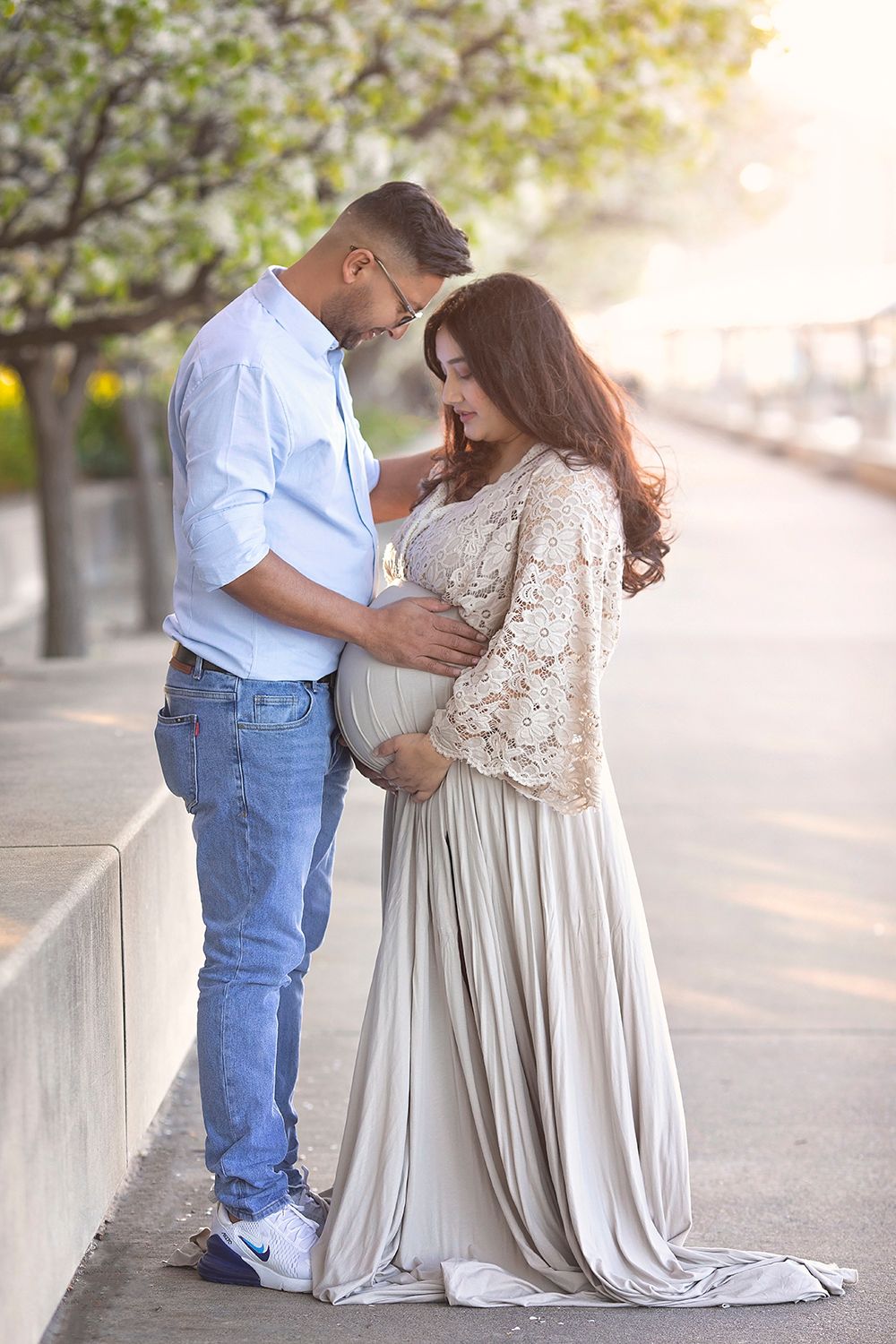 Couple shares tender moment outdoors, pregnant woman in lace and gray gown under blossoming trees.