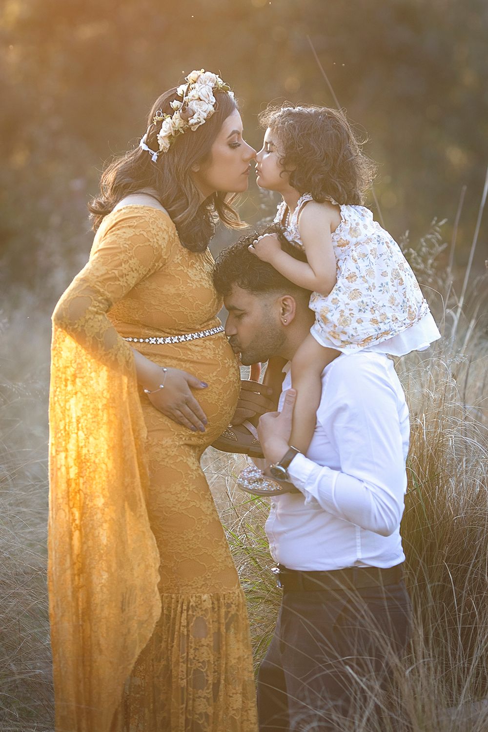 Pregnant woman in gold lace gown kisses toddler held by partner in sunlit golden field.