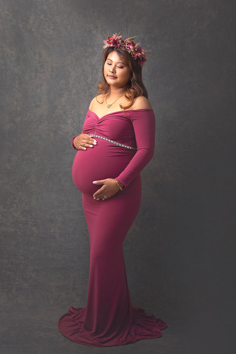Pregnant woman in elegant burgundy off-shoulder gown with floral crown posing for maternity photo.