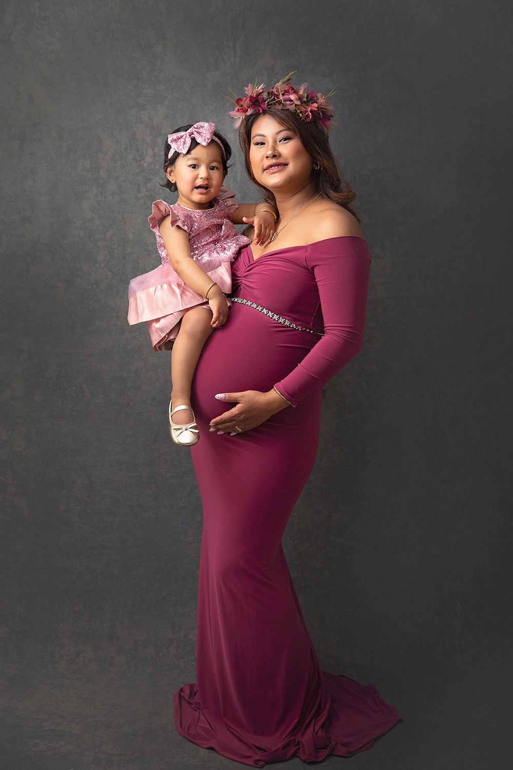 Pregnant woman in burgundy maternity gown holds toddler daughter in matching pink dress.