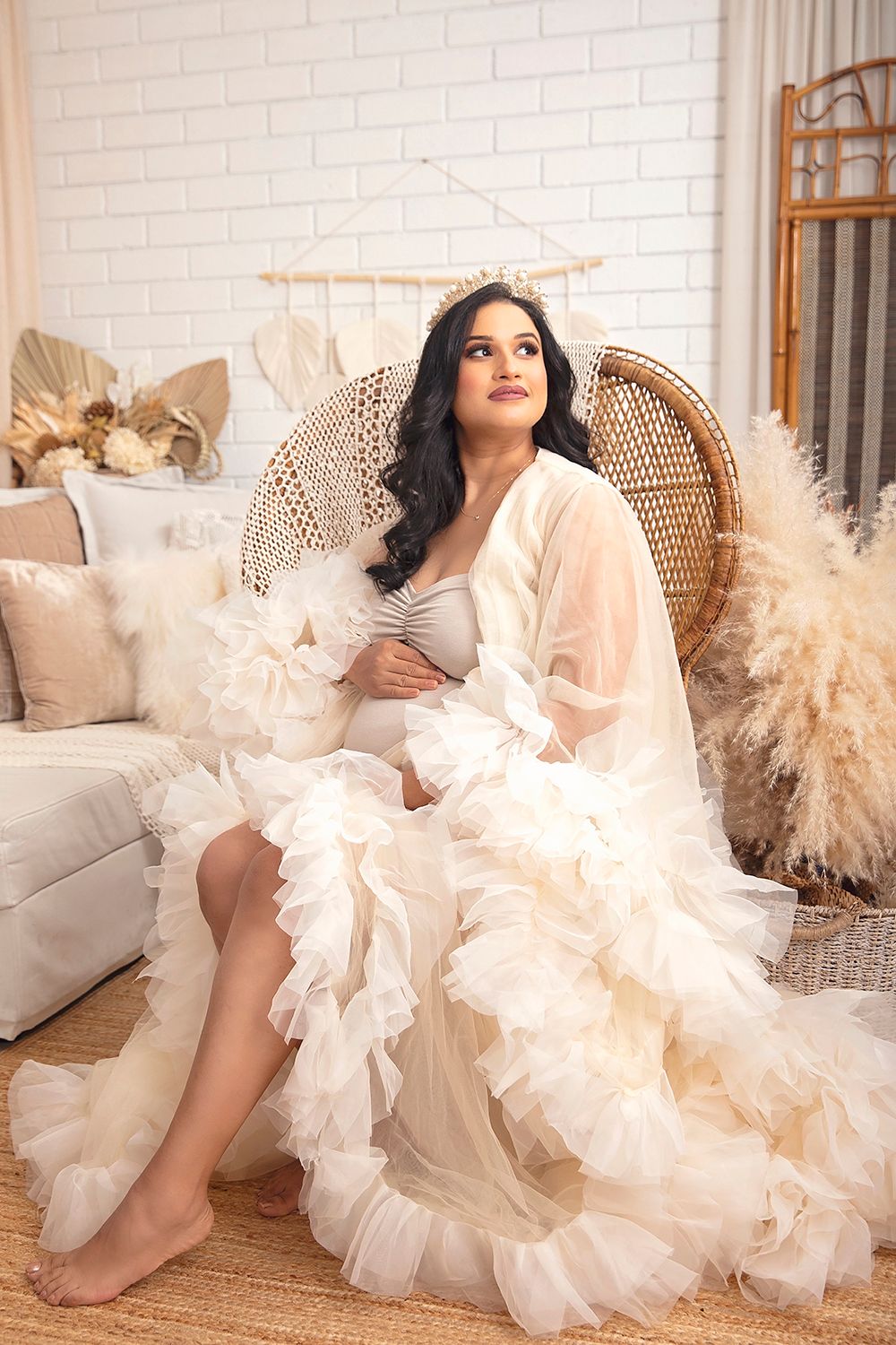 Pregnant woman in ivory ruffled tulle gown with crown sits in boho-styled studio setting.