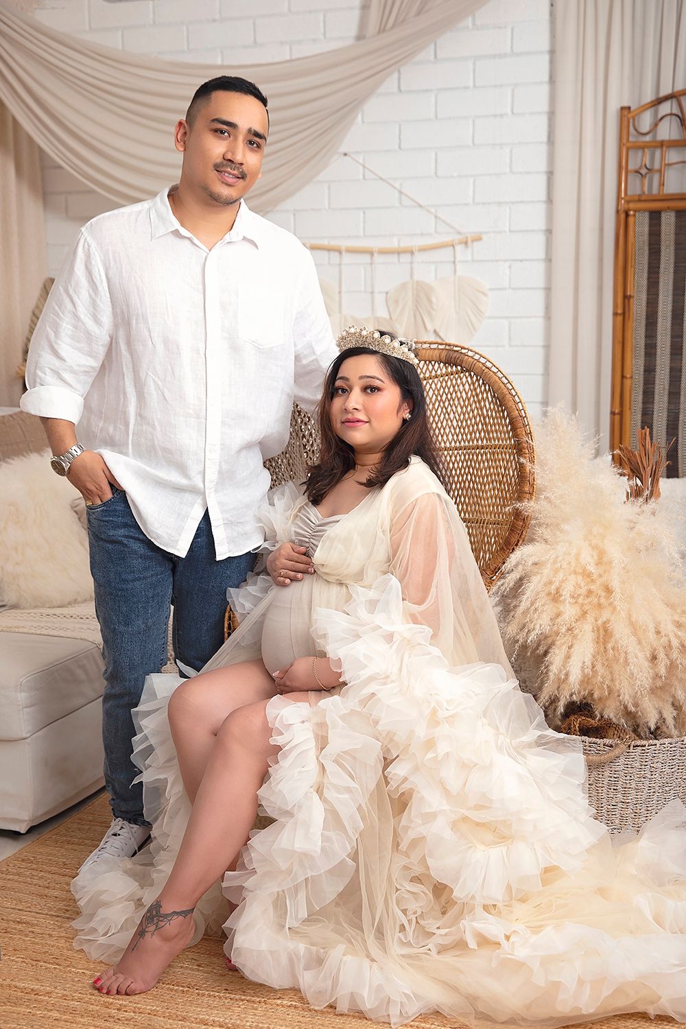 Expecting couple poses in boho studio, woman in cream ruffled gown, man in white shirt.