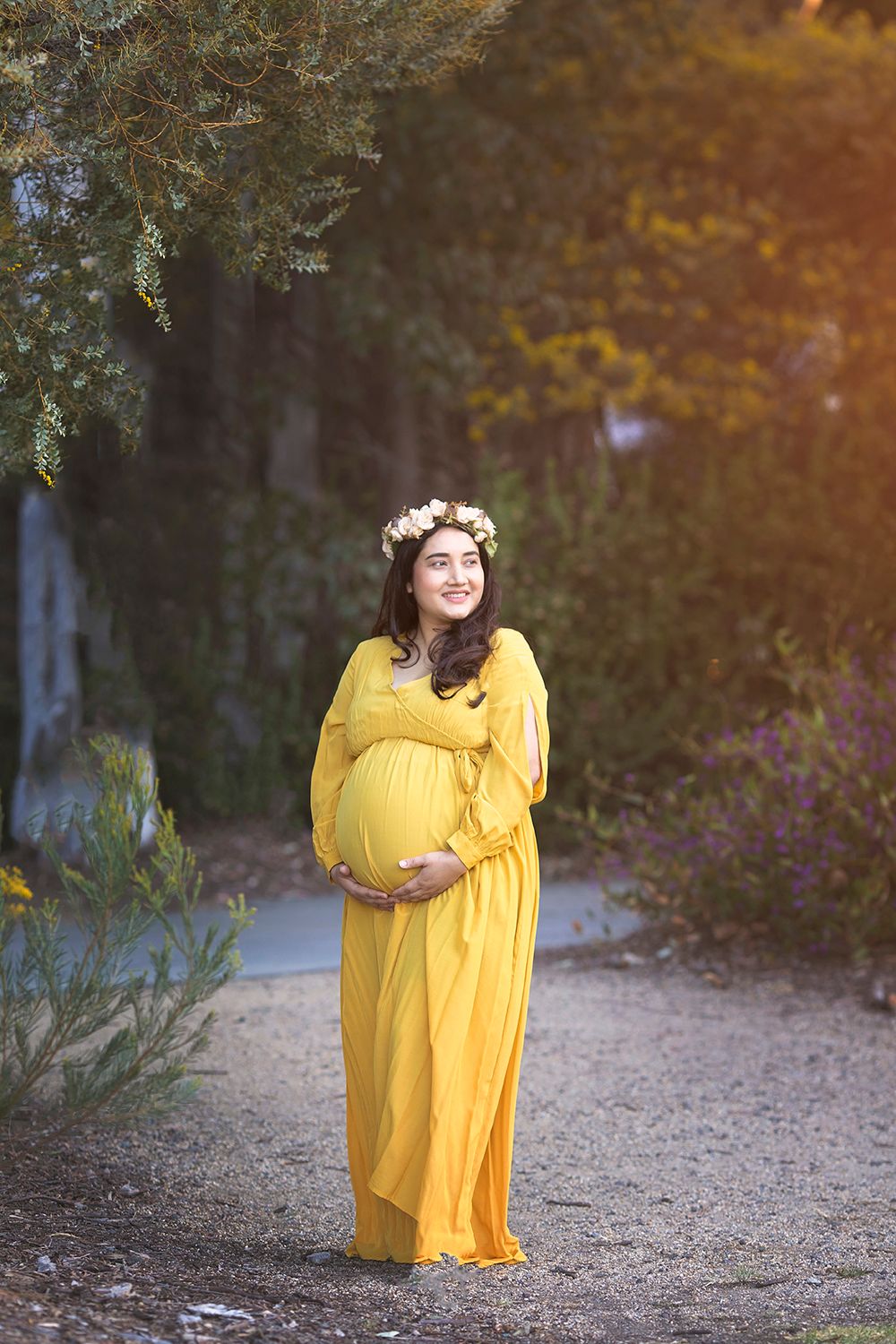 Pregnant woman in yellow off-shoulder gown with floral crown smiles outdoors in garden.