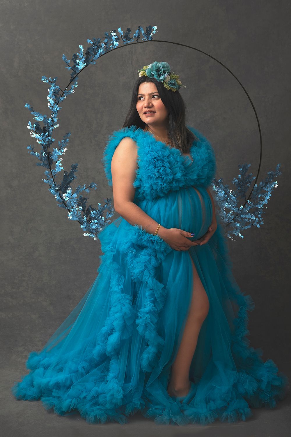 Pregnant woman in teal ruffled tulle gown poses before circular floral hoop backdrop.