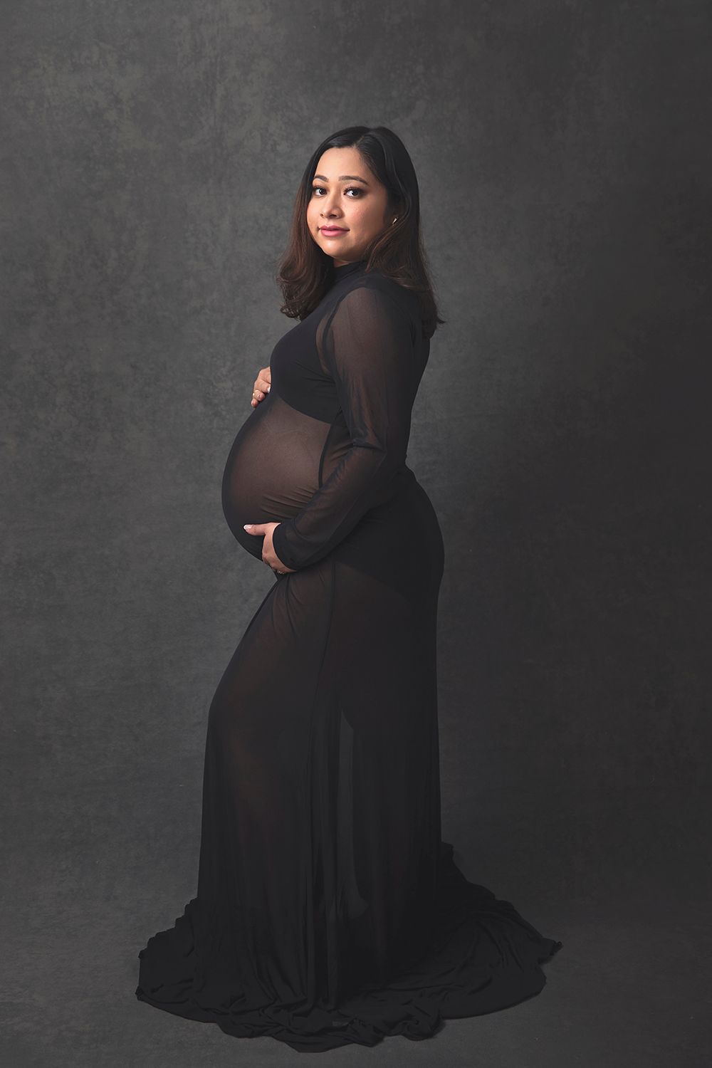 Pregnant woman in elegant sheer black maternity gown smiles over shoulder in studio portrait.