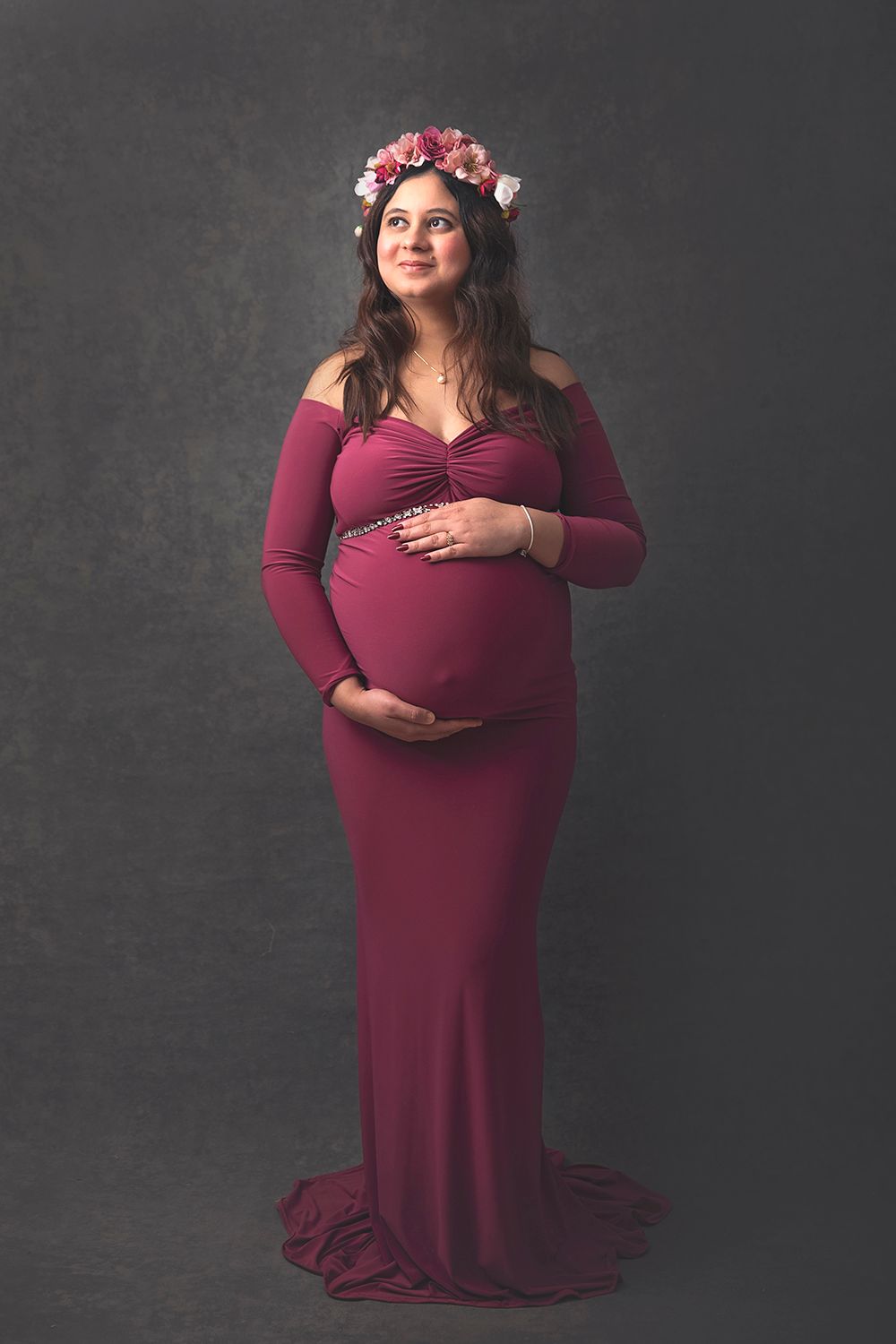 Smiling pregnant woman in burgundy off-shoulder gown with pink floral crown in studio.