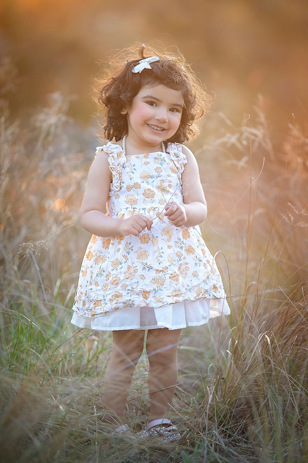 Smiling toddler girl in floral dress standing in golden field.