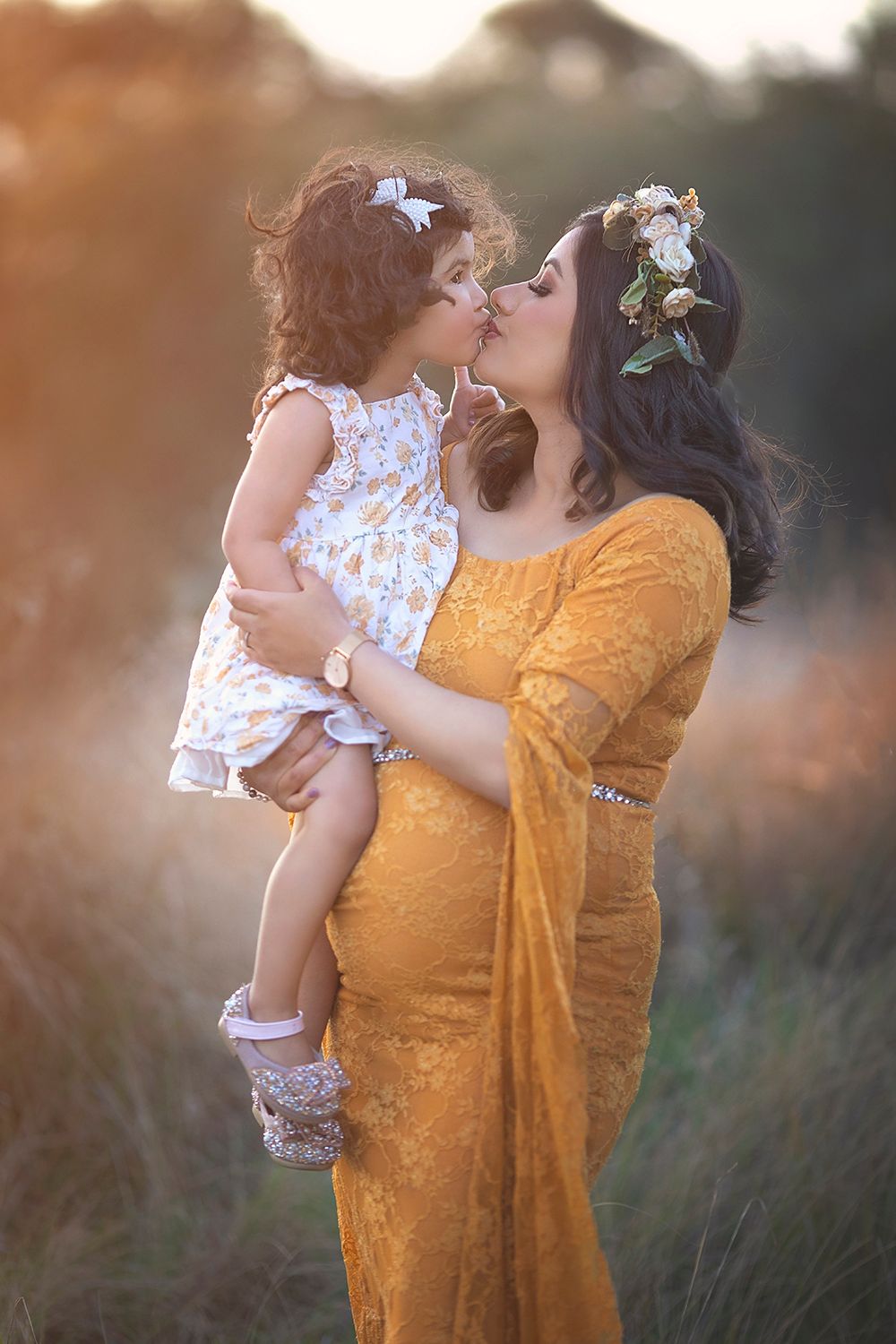 Pregnant woman in yellow lace gown kissing toddler daughter.