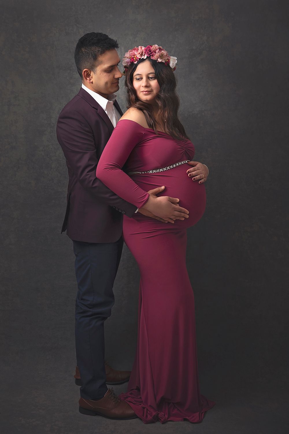 Couple maternity portrait in burgundy outfits on dark background.