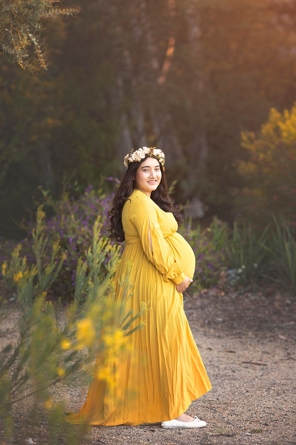 Pregnant woman in yellow maxi dress with floral crown outdoors.