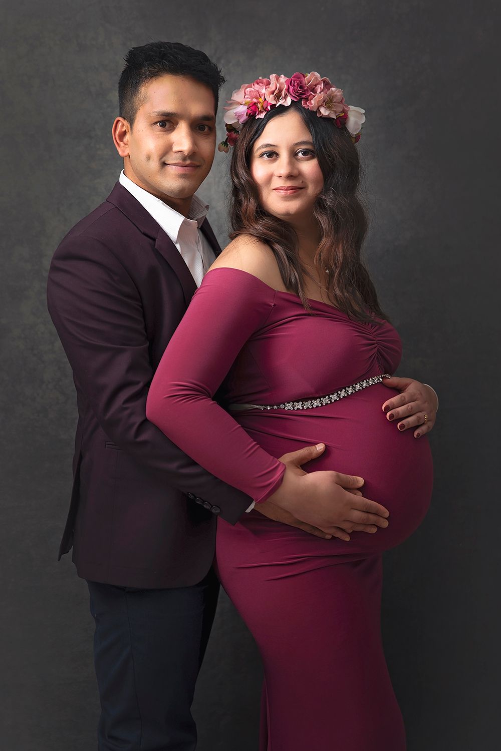 Couple maternity portrait in burgundy outfits on dark background.