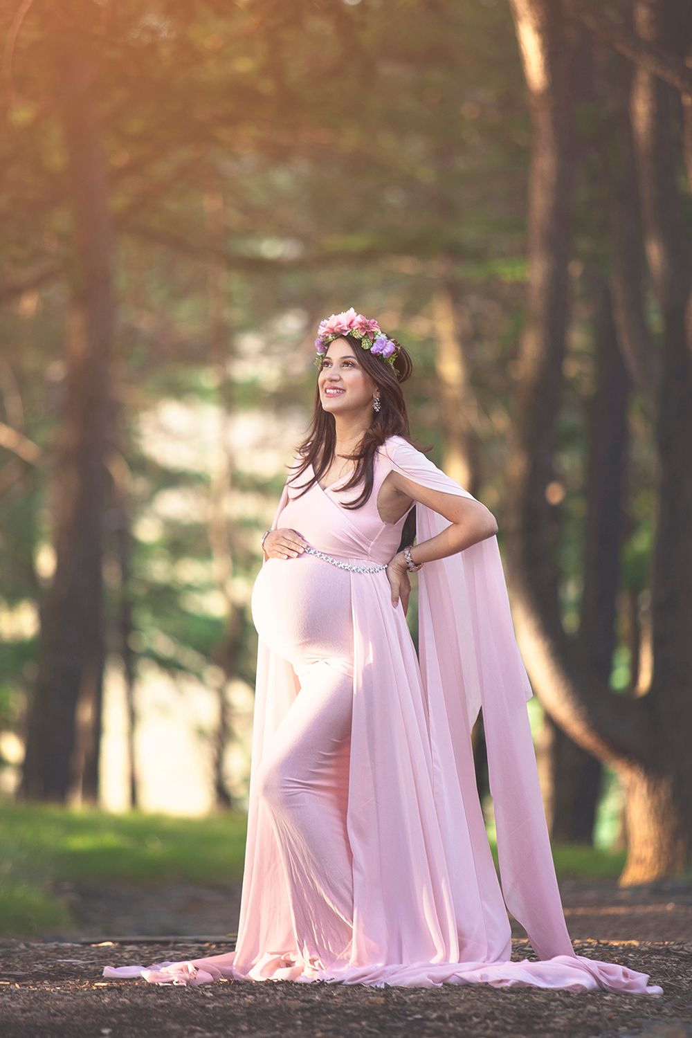 Pregnant woman in pink flowing gown with floral crown in forest.