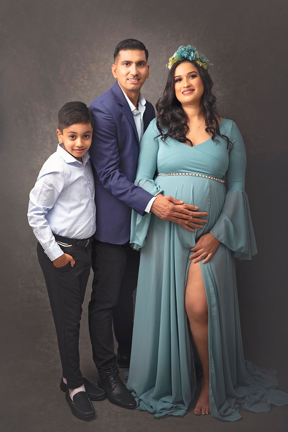 Family maternity photo with pregnant woman in teal gown, man in blue suit, and young boy in studio.