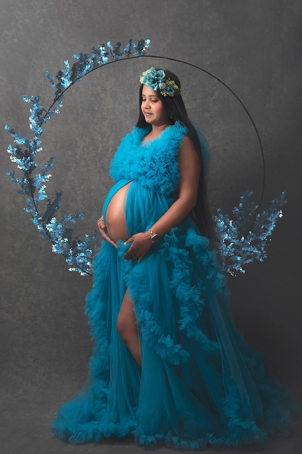 Pregnant woman in turquoise ruffled gown with floral crown poses before decorative blue floral hoop.