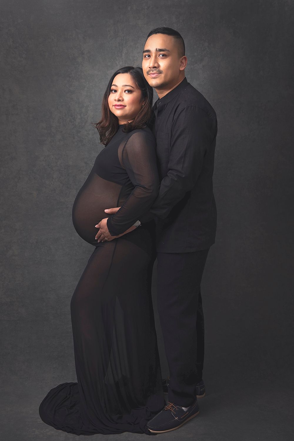 Couple in matching black outfits poses for maternity portrait in professional studio setting.