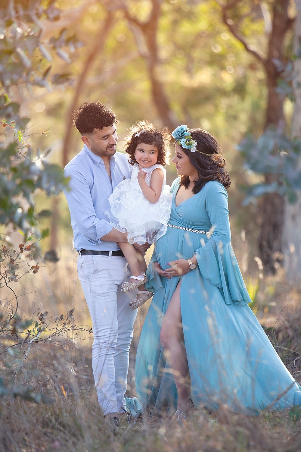 Family maternity outdoor portrait with pregnant woman in teal gown, man, and toddler in nature.