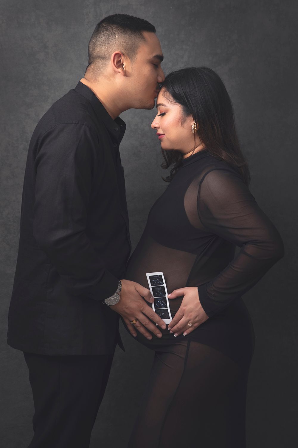 Couple in black outfits shares intimate maternity portrait with ultrasound photo in studio.