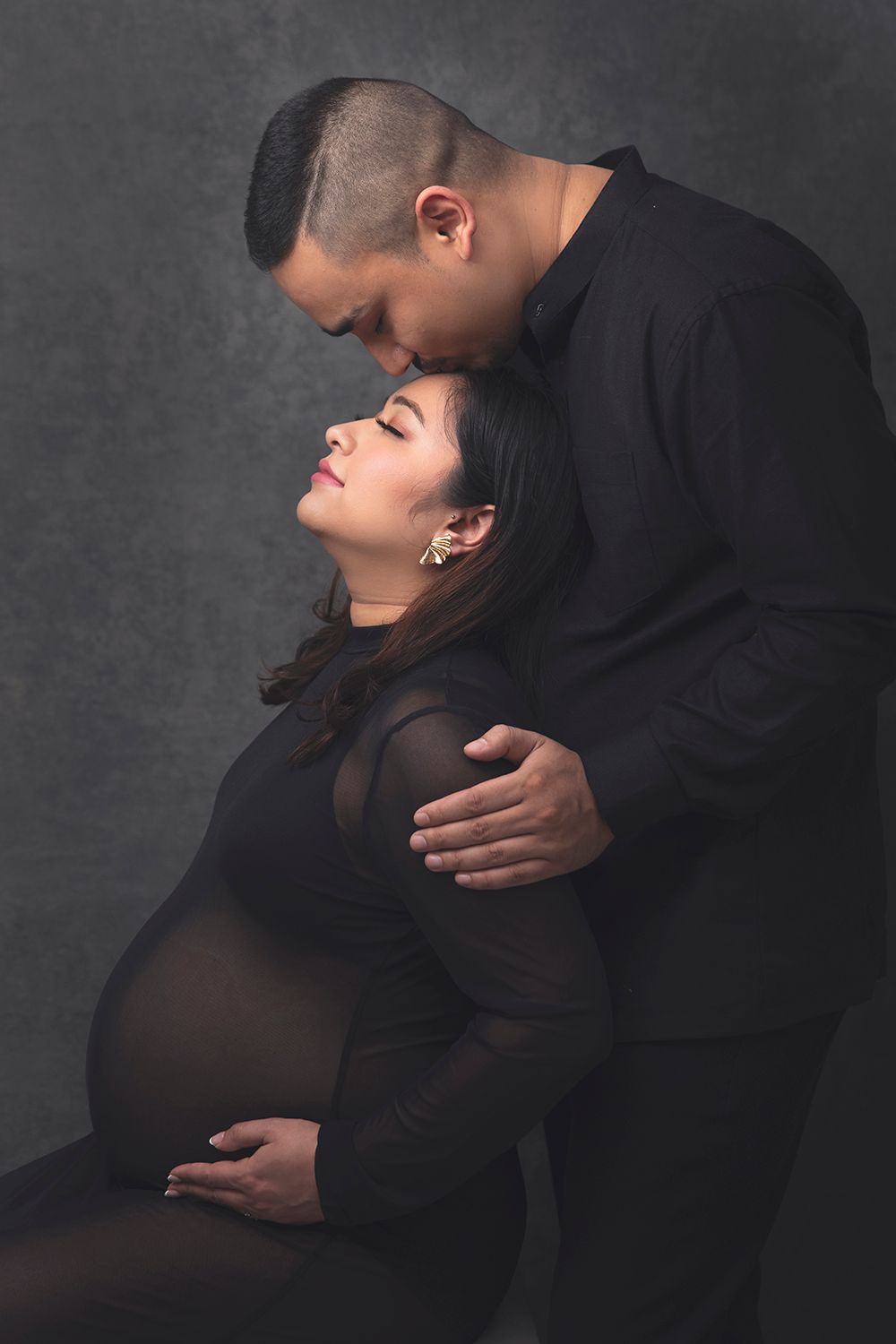 Intimate maternity photo of a pregnant woman in black dress with partner kissing her forehead against grey backdrop.