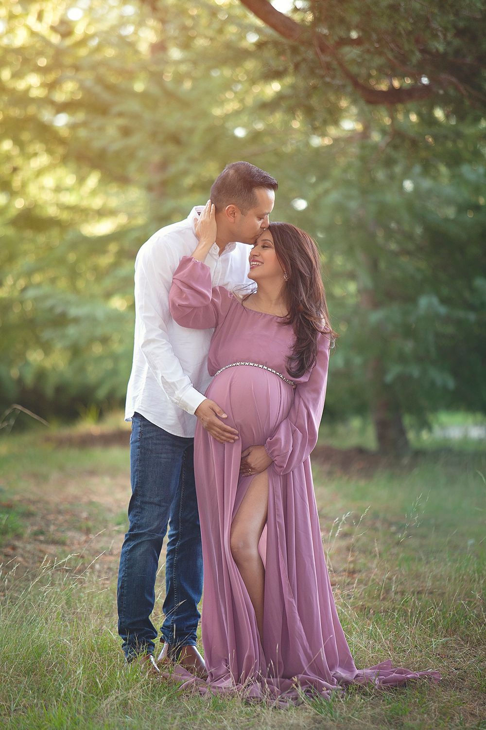 Couple embracing outdoors at golden hour, pregnant woman in flowing mauve gown during maternity photoshoot.